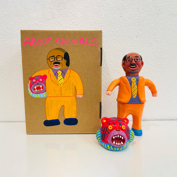 Sofubi figure of a Japanese business man in a bright orange suit and a yellow striped tie. At his feet is a pink tiger head, which can go over his own like a mascot head. It stands next to a painted box.