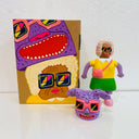 Sofubi figure of a woman in a bright yellow shirt and green skirt. At his feet is a comical purple bunny head with floppy ears and pink sunglasses, which can go over her own like a mascot head. It stands next to a painted box.