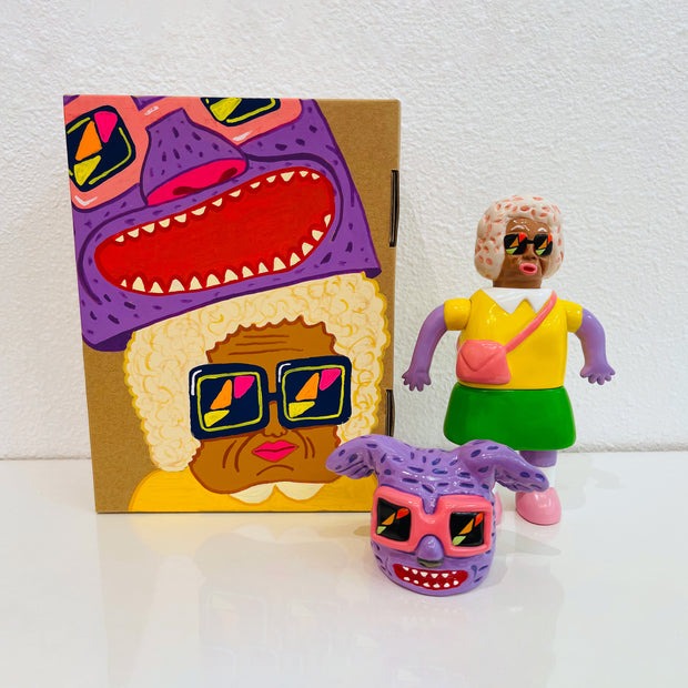 Sofubi figure of a woman in a bright yellow shirt and green skirt. At his feet is a comical purple bunny head with floppy ears and pink sunglasses, which can go over her own like a mascot head. It stands next to a painted box.
