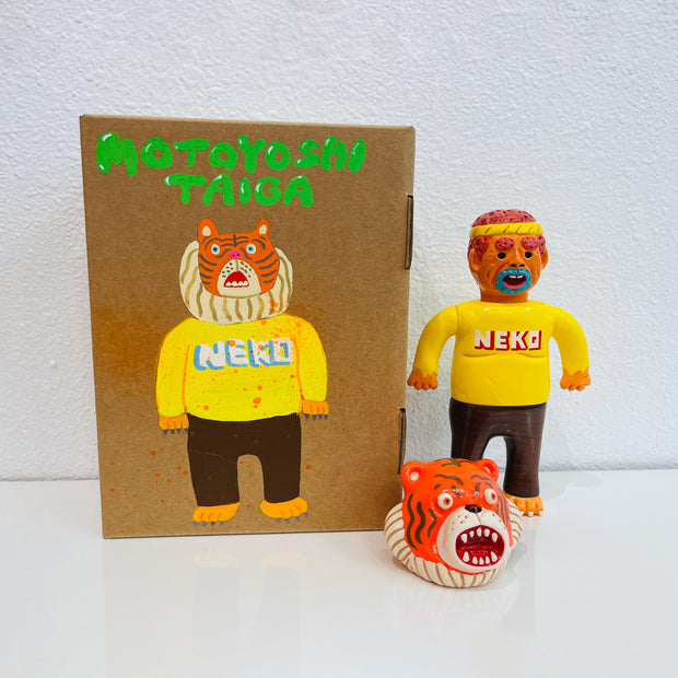Sofubi figure of a man in a bright yellow shirt that says "NEKO", brown pants and clawed feet. At his feet is an orange tiger head, which can go over his own like a mascot head. It stands next to a painted box.