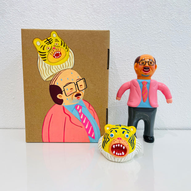 Sofubi figure of a Japanese business man in a bright pink blazer with grey slacks. At his feet is a yellow tiger head, which can go over her own like a mascot head. It stands next to a painted box.