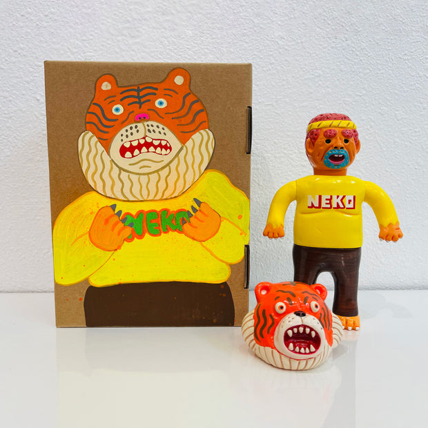 Sofubi figure of a man in a bright yellow shirt that says "NEKO", with brown pants and clawed feet. At his feet is an orange tiger head, which can go over his own like a mascot head. It stands next to a painted box.