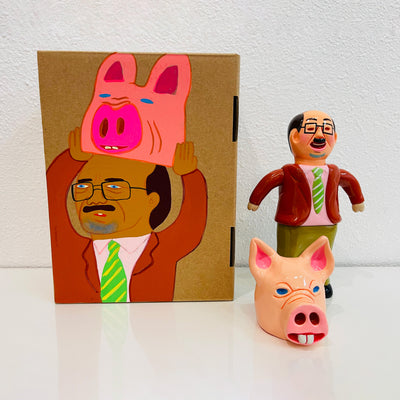 Sofubi figure of a Japanese business man in a burnt orange/red blazer with brown slacks. At his feet is a light pink pigs head, which can go over his own like a mascot head. It stands next to a painted box.