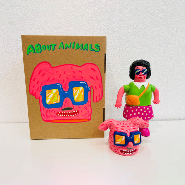 Sofubi figure of a woman with permed hair, glasses and brightly colored clothes. At her feet is a comical pink bunny head with blue sunglasses, which can go over her own like a mascot head. It stands next to a painted box.