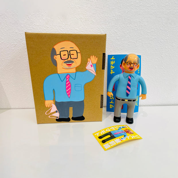 Soft vinyl figure of a business man wearing a light blue button up and grey slacks. He stands next to a painted box.
