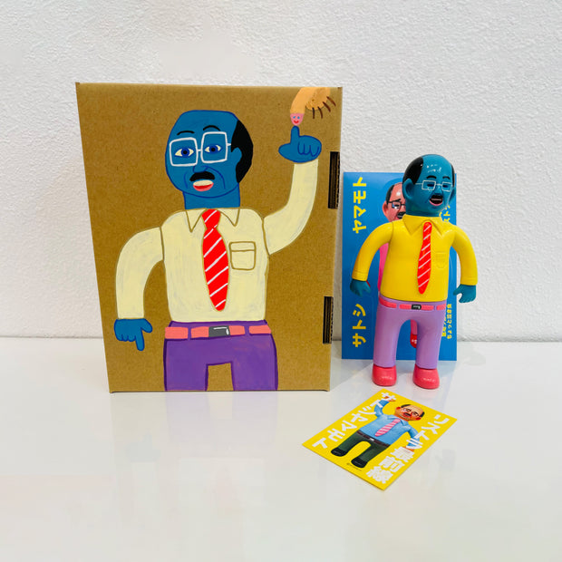 Soft vinyl figure of a blue business man wearing a bright yellow button up and purple slacks. He stands next to a painted box.