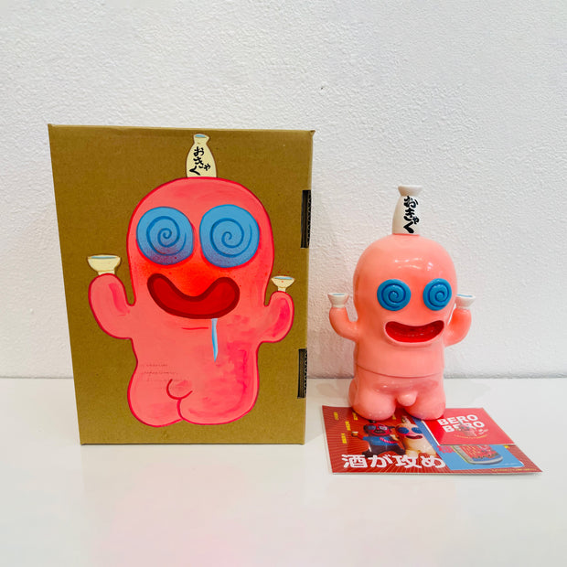 Light pink soft vinyl figure of a chubby character with a large, round head. It has a smiling face and swirled blue eyes. Atop its head is a bottle of sake, with a cup in each of its hands. It stands next to a painted box.
