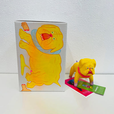 Yellow soft vinyl figure of a pitbull type dog, with a red collar and white paws. It stands in front of its product packaging with a few notecard underneath its box.