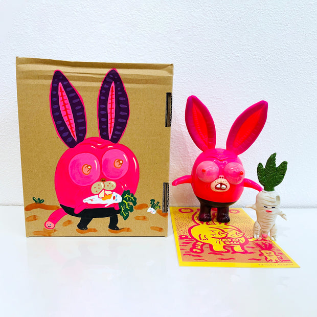 Neon pink vinyl figure of a strange looking rabbit, with female genitalia for ears, boobs for eyes and short body. It stands next to a short white daikon, with male genitalia. They stand in front of their product packaging.