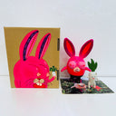 Neon pink vinyl figure of a strange looking rabbit, with female genitalia for ears, boobs for eyes and short body. It stands next to a short white daikon, with male genitalia. They stand in front of their product packaging.