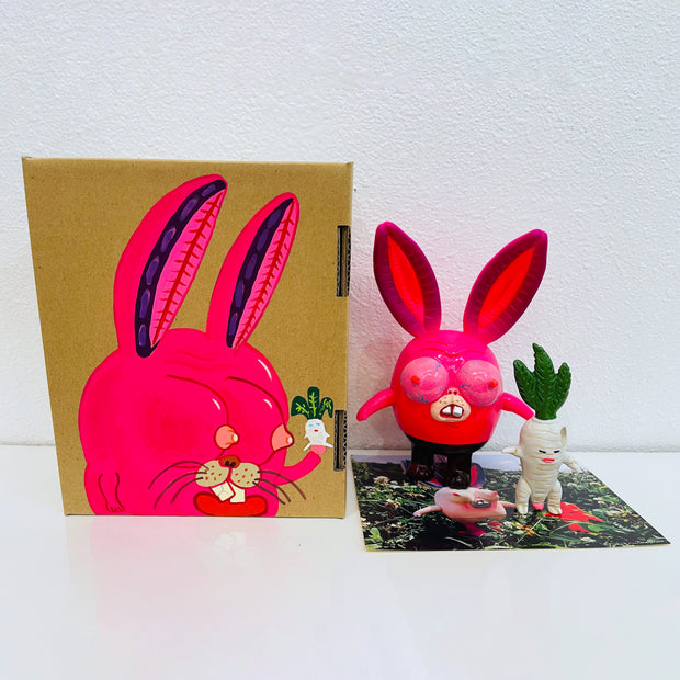Neon pink vinyl figure of a strange looking rabbit, with female genitalia for ears, boobs for eyes and short body. It stands next to a short white daikon, with male genitalia. They stand in front of their product packaging.