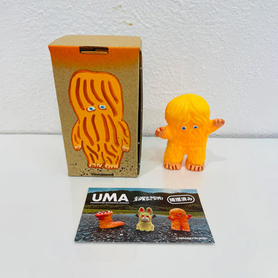 Small soft vinyl orange figure of a small monster with hair all over its body and 2 small eyes. It stands next to a painted box.