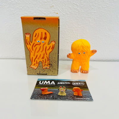 Small soft vinyl orange figure of a small monster with hair all over its body and 2 small eyes. It stands next to a painted box.