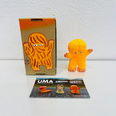 Small soft vinyl orange figure of a small monster with hair all over its body and 2 small eyes. It stands next to a painted box.