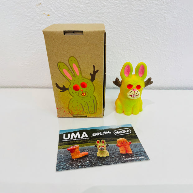 Small soft vinyl figure of a jackalope, bright yellow with neon pink eyes. It sits next to a painted box.