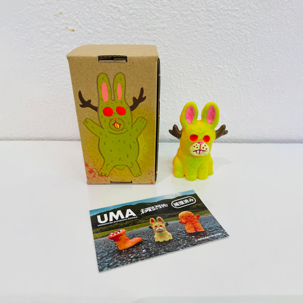 Small soft vinyl figure of a jackalope, bright yellow with neon pink eyes. It sits next to a painted box.