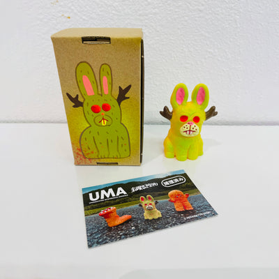 Small soft vinyl figure of a jackalope, bright yellow with neon pink eyes. It sits next to a painted box.