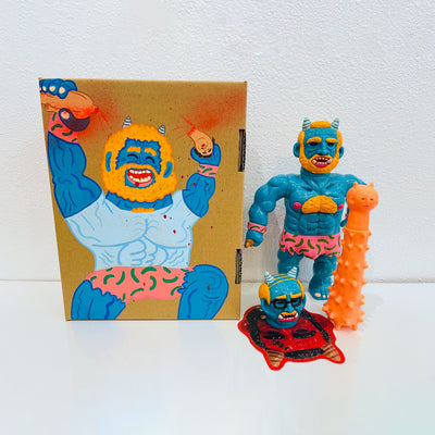 Blue soft vinyl figure of a demon man, with muscles and horns atop his head. He has a large orange spiked club and a human head at his feet. He stands next to a painted box.