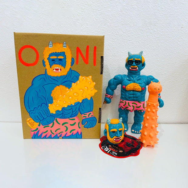 Blue soft vinyl figure of a demon man, with muscles and horns atop his head. He has a large orange spiked club and a human head at his feet. He stands next to a painted box.
