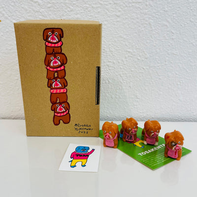 4 small, brown soft vinyl bulldogs with their mouths open and tongues out. They each have bright pink eyes and pink fabric around their neck, some with writing, and some blank. They stand in front of their product packaging.