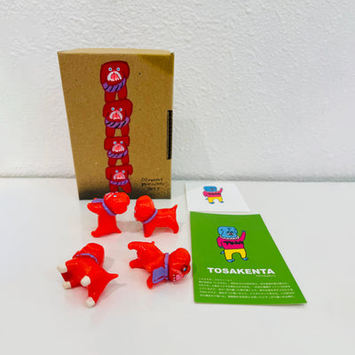 4 small, red soft vinyl bulldogs with their mouths open and tongues out. They each have bright pink eyes and purple fabric around their neck, some with writing, and some blank. They stand in front of their product packaging.