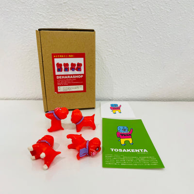 4 small, red soft vinyl bulldogs with their mouths open and tongues out. They each have bright pink eyes and purple fabric around their neck, some with writing, and some blank. They stand in front of their product packaging.