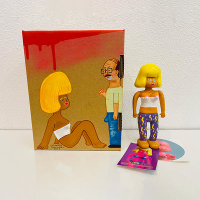 Tan soft vinyl figure of a woman in a white tube top and patterned pants, with large puffed blonde hair. She stands next to a painted box.