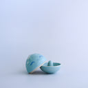 Small blue round bottomed jar with a lid. The lid has a slightly pointed tip wit white coloring and a drawn on, cute face. The lid is off and a ring holder is revealed on the inside.