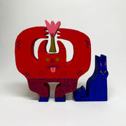 Wooden die cut sculptures of a red devil holding a flower over its head and a blue cat, sitting nearby. The devil has horns, green eyes, its tongue out and hairy goat legs.