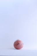 Small pink round bottomed jar with a lid. The lid has white coloring at the very top and a drawn on, cute face with rosy cheeks.