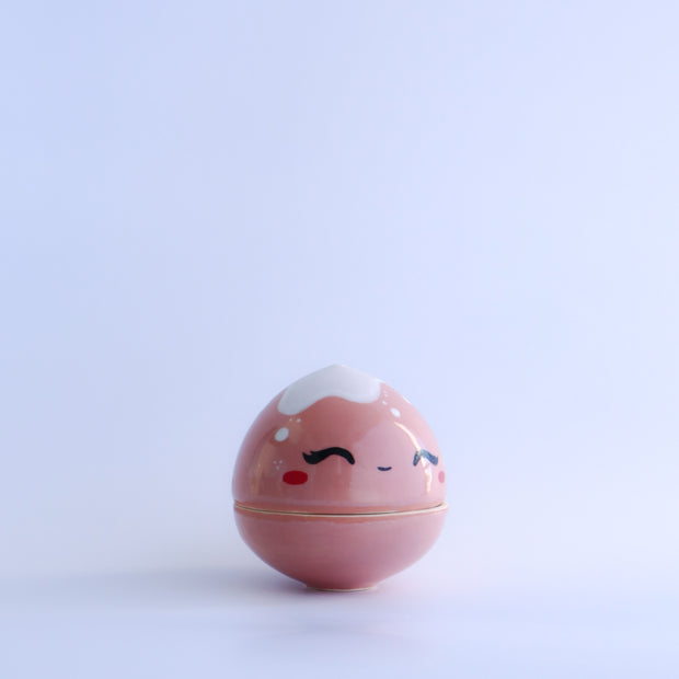 Small pink round bottomed jar with a lid. The lid has white coloring at the very top and a drawn on, cute face with rosy cheeks.