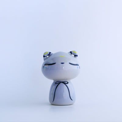Ceramic sculpture of a bluish gray cat, without any arms and legs. It wears a thin blue bow around its neck and has a calm, closed eye expression.