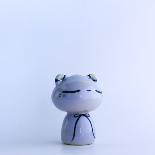 Ceramic sculpture of a bluish gray cat, without any arms and legs. It wears a thin blue bow around its neck and has a calm, closed eye expression.