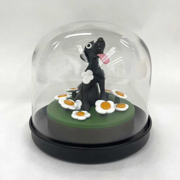 Sculpture made out of clay and under a clear dome of a black dog with its tongue out, on a mount of green ground with white daisies.