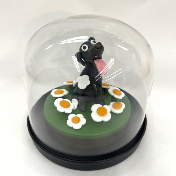 Sculpture made out of clay and under a clear dome of a black dog with its tongue out, on a mount of green ground with white daisies.