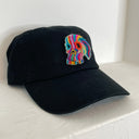 A black baseball cap featuring a colorful felted wool skull on the front, displayed against a plain white wall.