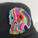 A black baseball cap featuring a colorful felted wool skull on the front, displayed against a plain white wall.