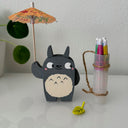 Small wooden sculpture of a smiling Totoro, looking off to the side. He holds up a small cocktail umbrella, with an over the shoulder bag holding 4 more umbrellas.
