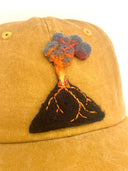 A close-up of a mustard yellow baseball cap featuring a hand-felted volcano patch on the front. The volcano has a black body, glowing orange lava lines, and gray and red smoke.