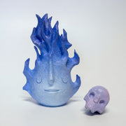 A small wooden sculpture of a purple skull and a taller sculpture of a blue flame with a closed eyed simplistic face.