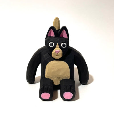 Whittled wooden sculpture of a fat black cat with pink ears and bottom of feet. It has a tan snout, belly and tail and looks straight at the viewer with its mouth slightly ajar, surprised looking.