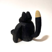 Whittled wooden sculpture of a fat black cat with pink ears and bottom of feet. It has a tan snout, belly and tail and looks straight at the viewer with its mouth slightly ajar, surprised looking.