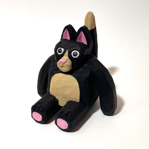 Whittled wooden sculpture of a fat black cat with pink ears and bottom of feet. It has a tan snout, belly and tail and looks straight at the viewer with its mouth slightly ajar, surprised looking.