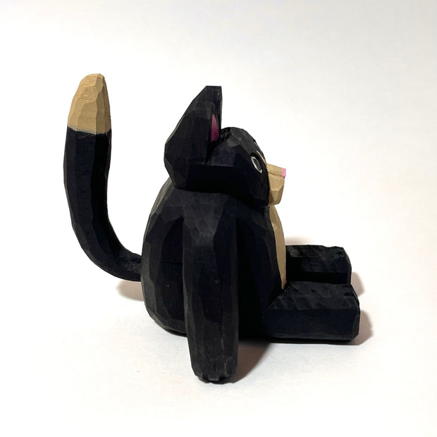 Whittled wooden sculpture of a fat black cat with pink ears and bottom of feet. It has a tan snout, belly and tail and looks straight at the viewer with its mouth slightly ajar, surprised looking.