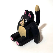 Whittled wooden sculpture of a fat black cat with pink ears and bottom of feet. It has a tan snout, belly and tail and looks straight at the viewer with its mouth slightly ajar, surprised looking.