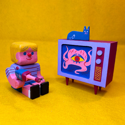 Colorful toy figure of a child watching a retro TV and holding a toy ray gun. Playing on the TV is a one eyed blob creature and a blue cat sits atop the TV. 