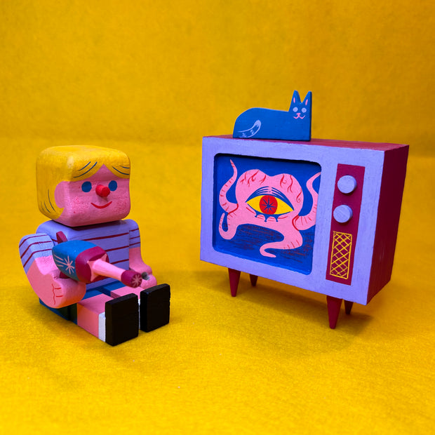 Colorful toy figure of a child watching a retro TV and holding a toy ray gun. Playing on the TV is a one eyed blob creature and a blue cat sits atop the TV. 