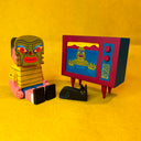 Colorful wooden sculpture of a boy sitting in front of a retro style TV. He wears a Loch Ness monster face mask, watching the Loch Ness monster on the TV next to a small black cat.