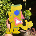 Die cut wooden sculpture of a Tiki man, an olive green with a large thumbs up.