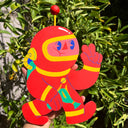 Colorful die cut wooden sculpture of a cartoon character with wearing a red space suit with yellow accents, held against a green leafy background.